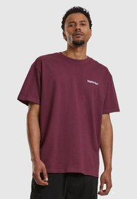 Tricou din bumbac burgund cu o croială lejeră, decolteu rotund și text alb "fețe uitate*" pe partea stângă a pieptului. Mâneci scurte.