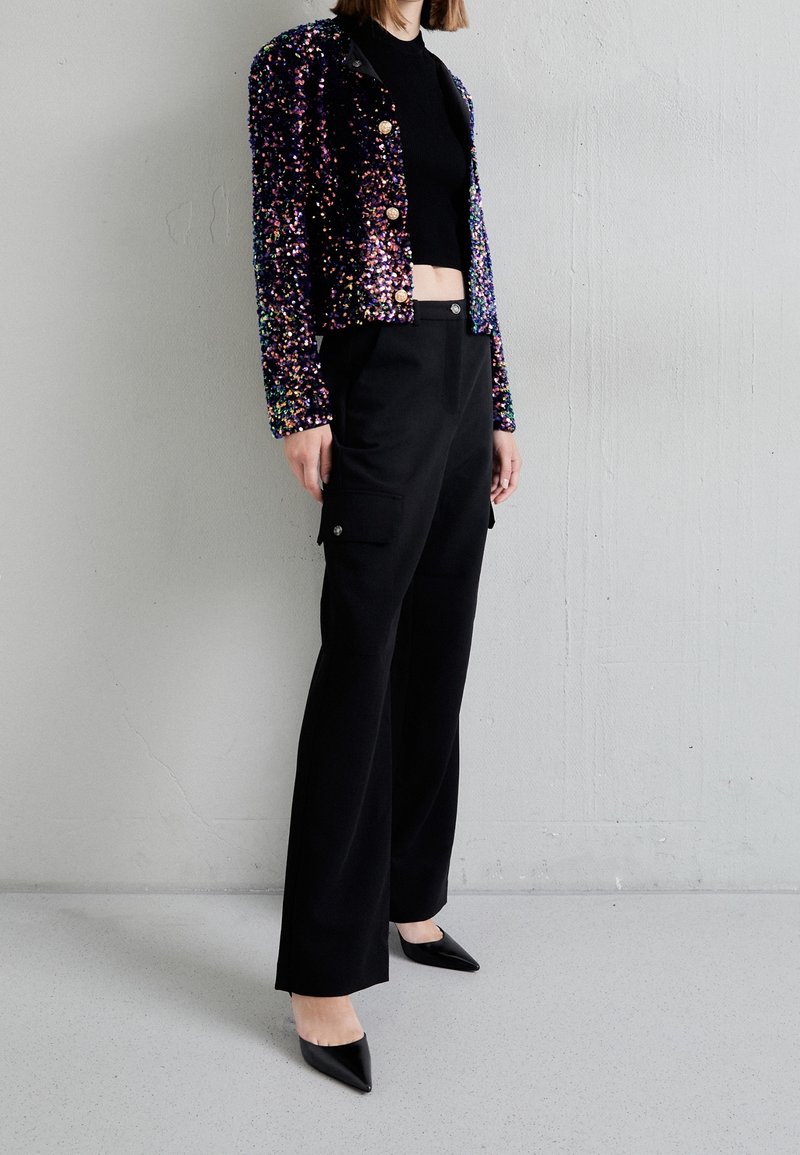 Personne portant une veste courte noire à sequins avec des boutons dorés, un pantalon noir taille haute, et des escarpins noirs à bout pointu, debout devant un mur gris.