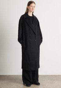 Abrigo negro de doble botonadura con grandes solapas, de corte oversize y bolsillos frontales. Combinado con pantalones negros holgados y zapatos negros.