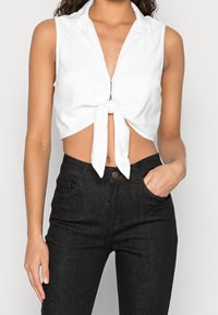 Top bianco accorciato senza maniche con colletto e dettaglio frontale annodato. Abbinato a jeans neri a vita alta con una texture liscia e una vestibilità classica.