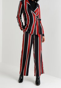 Ensemble tailleur rayé en noir, rouge et blanc ; composé d'un blazer ajusté avec un design enveloppant et d'un pantalon à jambes larges. Bottines noires brillantes.