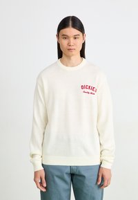 Pull en maille crème avec des poignets et un ourlet côtelés, mettant en avant un logo rouge brodé sur la poitrine gauche. Texture lisse et design décontracté.