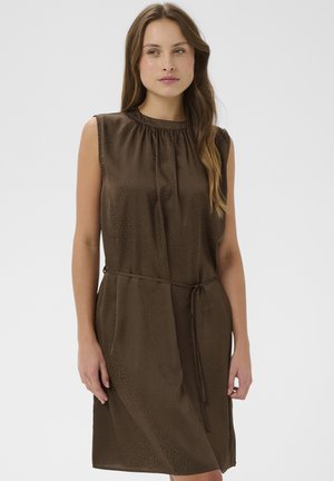 Žena s dlhými hnedými vlasmi, ktorá má na sebe tmavohnedé šaty bez rukávov dĺžky po kolená s úzkym opaskom zviazaným v páse, stojaca na neutrálnej pozadí.