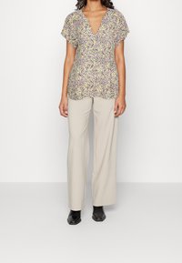 Blouse à manches courtes florale en blanc, jaune et violet avec un décolleté en V ; associée à un pantalon large beige clair et des bottines noires.