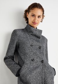Manteau en mélange de laine gris avec un col haut, une fermeture double boutonnage et des boutons noirs. Il présente un tissu texturé et des poches latérales.