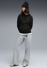 Camisola com capuz preta com logótipo, camada base branca, jeans azul claro de perna larga e ténis bege. Modelo a usar um gorro preto, em pé com os braços cruzados.