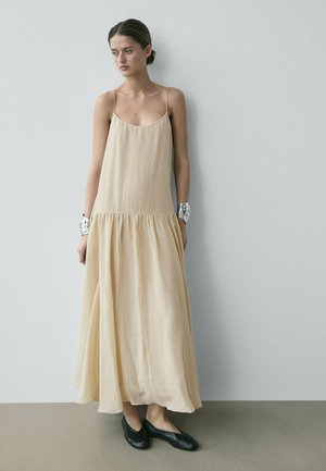 Donna che indossa un vestito lungo beige senza maniche con spalline sottili, bracciali a polsino argentati e scarpe basse nere, in piedi contro un muro semplice.