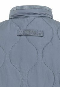 Graue gesteppte Jacke hinten mit gebogenen Nahtmustern und einem Stoffetikett mit der Aufschrift "camel active", das unter dem Kragen eingenäht ist.