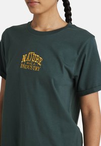 Dark green short-sleeve t-shirt with embroidered "NATURE RVCA INDUSTRY" in yellow. Round neck and a relaxed fit. Soft cotton material.