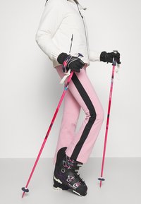 Lyžařský outfit zahrnující bílou bundu, růžové kalhoty s černým bočním pruhem, černé lyžařské boty a růžové lyžařské hole, všechno v dynamické lyžařské pozici.