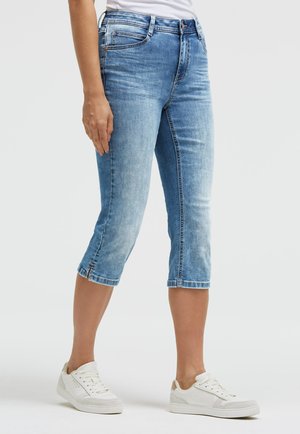 Woman wearing light blue denim capri jeans and white sneakers standing against a plain background.