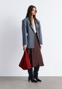 Grauer Wollblazer, cremefarbene, knöpfbare Bluse, brauner Satinsrock, roter Handtasche, schwarze kniehohe Stiefel mit strukturiertem Finish. Minimalistische Umgebung.