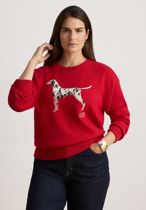 Mujer con el pelo largo y castaño que lleva un suéter rojo con un diseño de perro dálmata, vaqueros anchos oscuros, zapatos negros y una pulsera de plata.