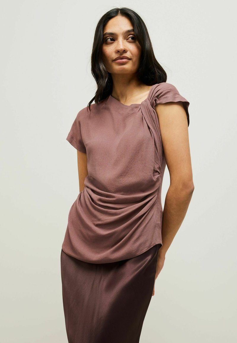 Top in tessuto malva con maniche corte, caratterizzato da dettagli drappeggiati su un lato e una texture liscia. Abbinato a una gonna elegante e coordinata.