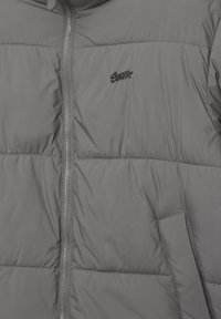 Szürke steppelt dzseki elülső cipzárral és egy kis hímzett logóval a bal mellrészen, egy oldalzseb felett.