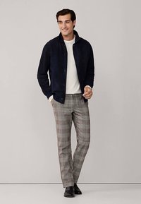 Veste en daim marine portée sur un pull blanc, associée à un pantalon à carreaux gris. La tenue se caractérise par un design à boutons et des chaussures noires unies.
