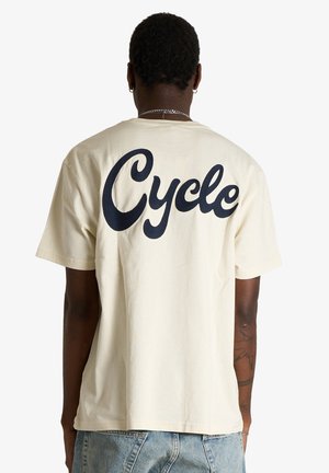 Persona che indossa una camicia beige chiaro a maniche corte con grande scritta nera "Cycle" sulla schiena, abbinata a jeans azzurri, vista di spalle.