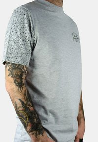 Camiseta gris de manga corta con un patrón de triángulos en la manga negra, cuello redondo, material de algodón y un gráfico geométrico minimalista en el pecho.