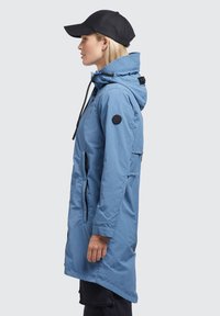 Parka impermeabilă de culoare albastru deschis, cu glugă cu șnur, buzunare laterale, fermoare negre contrastante și o pată circulară cu logo pe brațul superior.