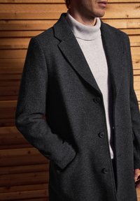 Sivý vlněný kabát s klopou, dvěma knoflíky a bočními kapsami, připevněný na světle šedém roláku. Texturovaná látka.