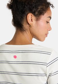 Frau mit lockigem Haar zu einem Dutt frisiert, trägt goldene Creolen und ein weißes, geripptes Shirt mit schwarzen Streifen und einem kleinen rosa runden Logo auf dem Rücken.