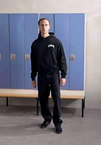 Black hooded sweatshirt with a front pocket and logo, paired with black jeans and dark sneakers, set against blue lockers.