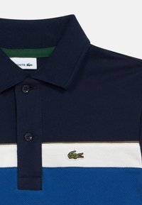 Moška polo majica, mornarsko modra z zelenim ovratnikom, ima vodoravne črte v beli in modri barvi, teksturiran material in majhen krokodilov logotip.