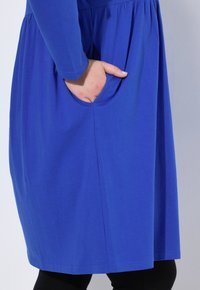 Modré šaty vyrobené z měkké látky, s dlouhými rukávy a staženým pasem, s kapsou na boku. Hladká textura a jednoduchý design.