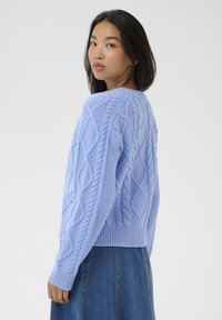 Pull en tricot bleu clair avec un motif de losanges texturé, des poignets et un ourlet côtelés. La vue arrière révèle des détails de tricot en câble complexes.