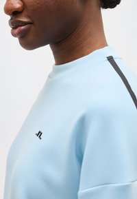 Detailný záber osoby, ktorá má na sebe svetlomodrú mikinu s malým čiernym logom a čiernym pruhom na ramene.