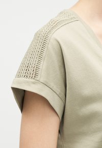 Top beige de manga corta con cuello en V y detalle de patrón de punto intrincado que recorre la costura del hombro.