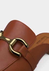 Mule en cuir marron avec bride texturée et détail boucle en métal doré en forme de mors de cheval sur le dessus.