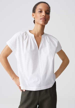 Femme portant un chemisier blanc à manches courtes avec un petit col en V et un pantalon noir, debout les mains sur les hanches sur un fond uni.