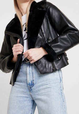 Femme portant une veste en cuir noire avec doublure en fourrure, zippée à mi-hauteur, associée à un jean taille haute bleu clair et un t-shirt blanc à motif.