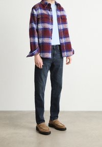 Hombre vestido con una chaqueta de cuadros en color marrón, azul y blanco, sobre una camisa azul clara, jeans oscuros y mocasines en color beige, de pie sobre un suelo de concreto.