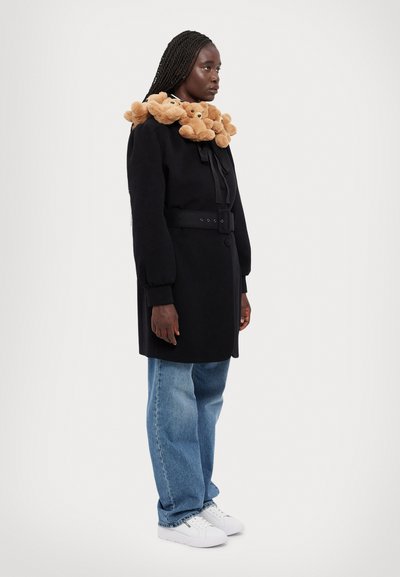 Zwarte jas met pluizige teddybeeraccenten rond de kraag, een ceintuur in de taille, lange mouwen, gecombineerd met lichtblauwe jeans en witte sneakers.