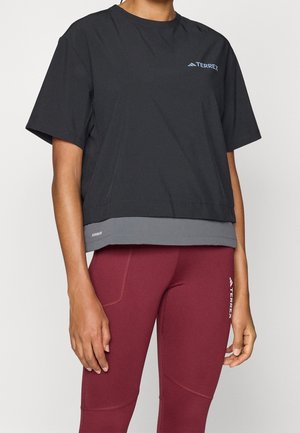 Osoba ubrana w czarną krótką koszulkę Terrex na szarym spodku, w połączeniu z bordowymi legginsami Terrex, widziana od ramion do połowy ud.