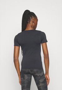 Tricou negru, ajustat, cu mânecă scurtă și textură netedă, cu gât rotund, purtat împreună cu colanți de camuflaj în nuanțe întunecate, văzut din spate.