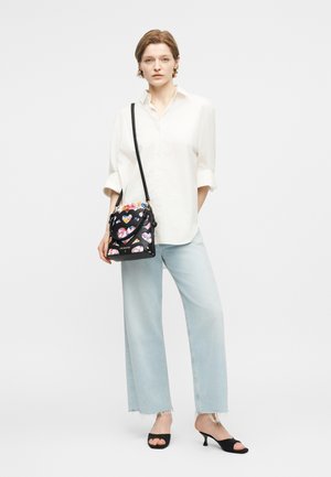 Γυναίκα που φοράει λευκό πουκάμισο με κουμπιά, ανοιχτό μπλε τζιν παντελόνι φαρδιάς γραμμής, μαύρες σανδάλες με τακούνι, και κρατάει μαύρη τσάντα χιαστί με πολύχρωμες καρδιές κομμένες.