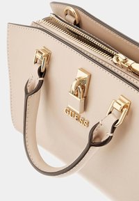 Sac à main en cuir synthétique beige avec quincaillerie dorée, double fermeture éclair et une courte poignée. Comprend un logo et des détails de couture contrastants.