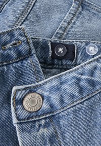 Ljusblå denimtyg med synliga sömmönster. Har en metallknapp och en klar plastknapp som stängning vid midjan.