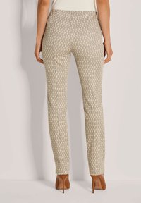 Vrouw in beige, met een patroon, slim fit broek en bruine hoge hakken, staand tegen een lichte muur, gezien van achteren.