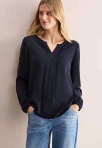 Mörkblå blus med V-ringning, lång ärm och elastiska manschetter. Tillverkad av lätt material med subtil textur och två vertikala sömmar.
