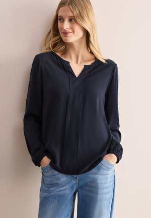Blouse bleu marine avec un col en V, des manches longues et des poignets élastiques. Fabriquée en tissu léger avec une texture subtile et deux coutures verticales.