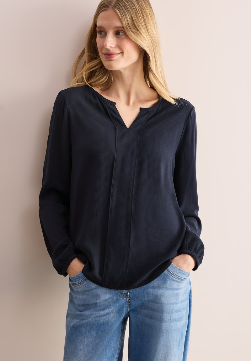 Mörkblå blus med V-ringning, lång ärm och elastiska manschetter. Tillverkad av lätt material med subtil textur och två vertikala sömmar.