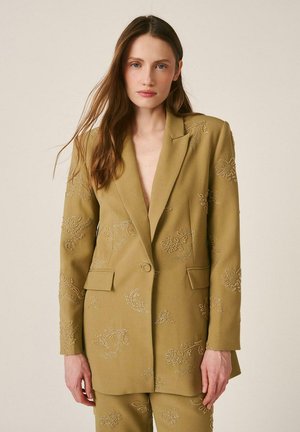 Femme aux longs cheveux bruns portant un blazer vert olive et un pantalon assorti avec une broderie florale subtile, debout devant un fond uni.