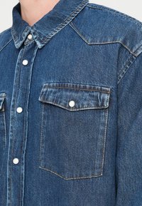 Μπλε πουκάμισο από τζιν με κουμπιά στο μπροστινό μέρος, δύο τσέπες στο στήθος και ασημί κουμπώματα snap. Διαθέτει δομημένο γιακά και ορατές διακοσμητικές ραφές.
