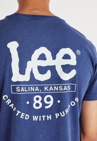 Niebieska bawełniana koszulka z dużym białym napisem na plecach: "Lee," "Salina, Kansas," "89," oraz "Crafted with Purpose." Prosty design.