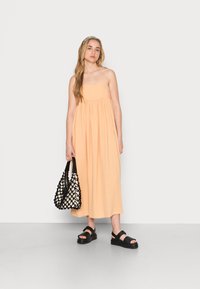 Vestido maxi sem mangas de cor pêssego com um design fluido. Bolsa tecida preta na mão e sandálias pretas grossas nos pés. Textura suave.