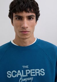 Jeune homme avec des cheveux courts et bouclés de couleur foncée portant un sweat-shirt bleu avec le texte "The Scalpers Company" au devant, fond neutre.